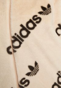 Tessuto peloso color crema con motivo ripetuto del logo Adidas nero e del trifoglio, separati da una cucitura verticale.