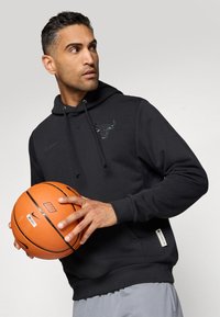 Svart hettegenser med et grafisk okse-logo, snorstrammet og kengurulomme. Modellen holder en oransje basketball og har på seg grå shorts.