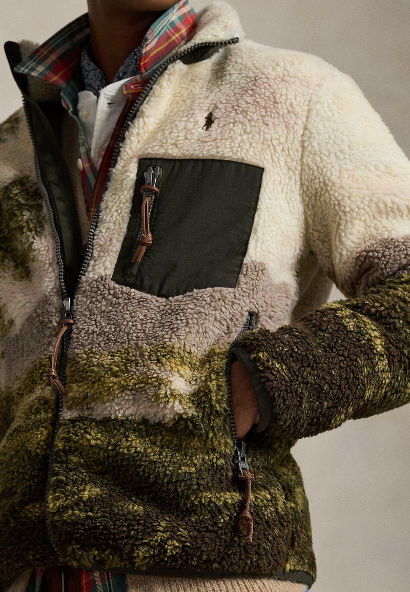 Giacca in pile con una combinazione di colori crema, oliva e marrone, con una tasca sul petto verde, dettagli in pelle e chiusure con cerniera.