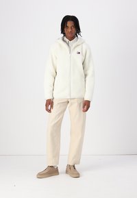Hvid fleece zip-up jakke med en tekstureret finish, beige bukser og lysebrune sko. Jakken har et lille logo.