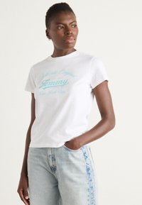 Hvit bomulls T-skjorte med turkise bokstaver som leser "Authentic Original siden 1983 Tommy fra New York City." Korte ermer, avslappet passform.