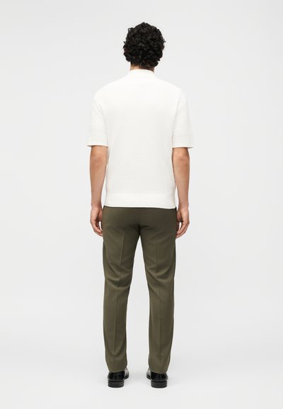 Άνδρας με σγουρά μαλλιά στέκεται με την πλάτη γυρισμένη, φοράει λευκό κοντομάνικο πουκάμισο, ελαιδί παντελόνια και μαύρα παπούτσια σε ένα μονόχρωμο φόντο.