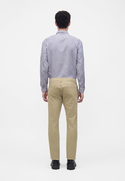 Homme portant un pantalon beige, des chaussures noires et une chemise rayée bleu et blanc à manches longues, debout de dos sur un fond blanc uni.