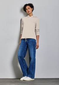 Sweat-shirt à capuche gris clair avec cordon de serrage, associé à un jean bleu ample et des baskets blanches, sur fond uni.