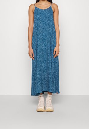 Blauwe, mouwloze maxi-jurk met een stippenpatroon, een relaxte pasvorm en een zijlabel. Gecombineerd met lichtroze sneakers.