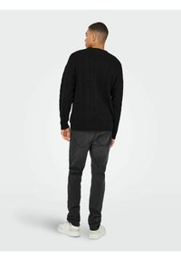 Schwarzer Strickpullover mit strukturiertem Muster, kombiniert mit dunkelgrauen Jeans und weißen Turnschuhen. Die Ansicht ist von hinten.