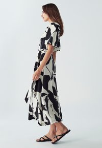 Schwarz-weiß gemustertes, knielanges Kleid mit kurzen Ärmeln und tailliertem Gürtel, kombiniert mit schwarzen Flip-Flops vor einem schlichten Hintergrund.