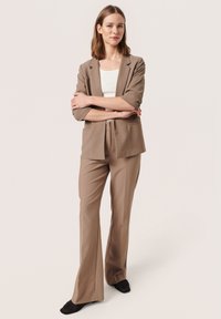 Hnědý tailored blazer a odpovídající rozšířené kalhoty vyrobené z hladkého materiálu. Nosit se může přes bílý top, má boční kapsy a přiléhavou siluetu.