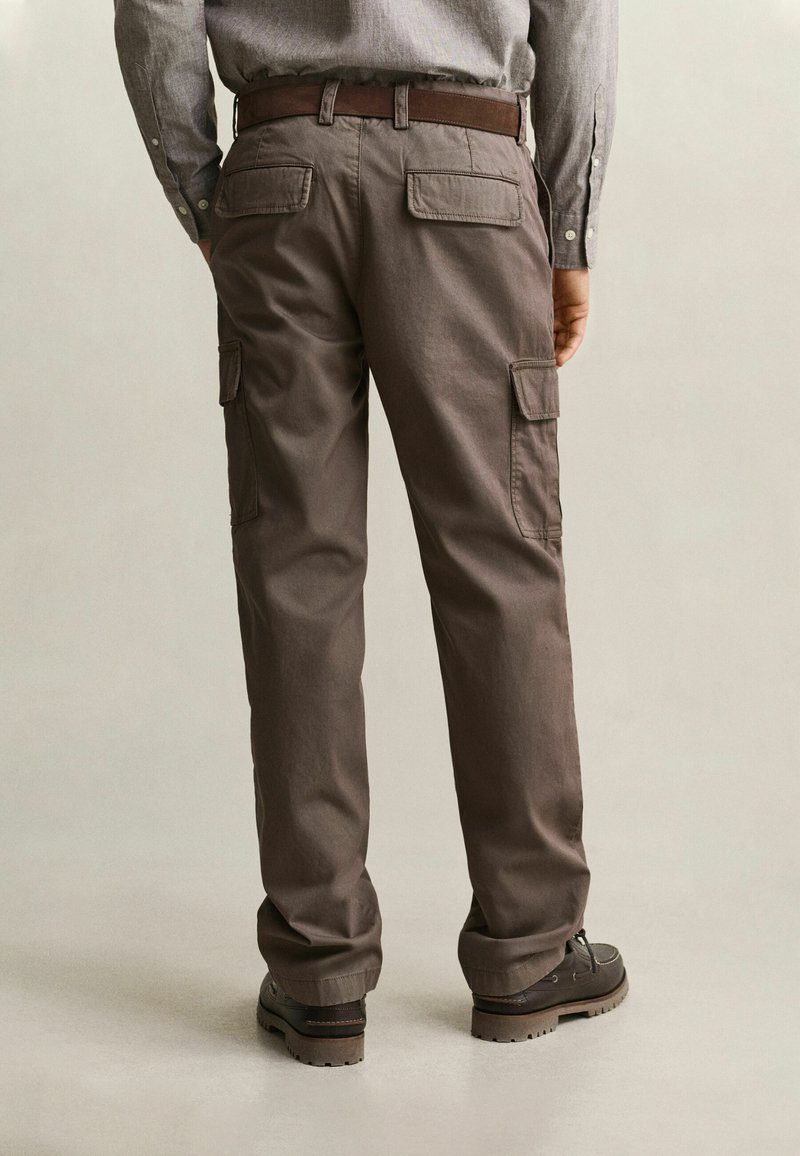 Hombre con pantalones cargo marrones, cinturón marrón oscuro, camisa gris de manga larga y zapatos marrón oscuro, de pie sobre un fondo claro de espaldas.