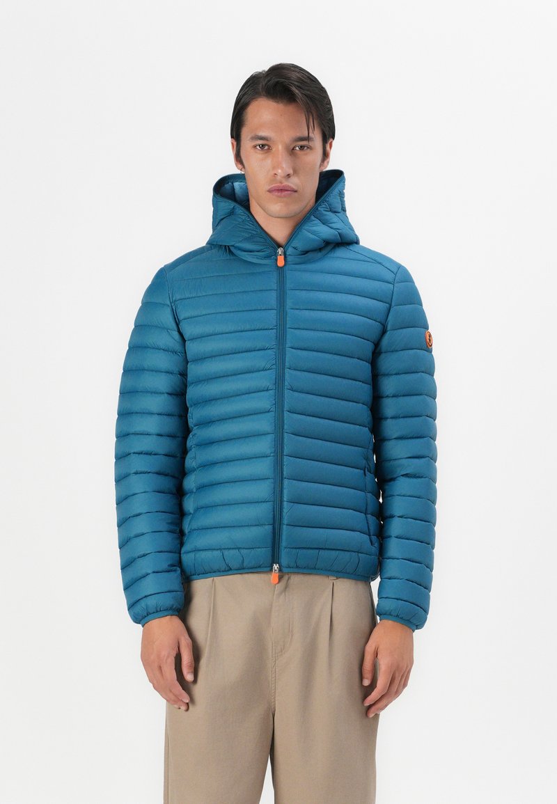 Geacă puffer teal cu glugă, cu un model cusut orizontal, fermoar frontal cu un tiv portocaliu și o aplicație cu logo pe mâneca stângă.