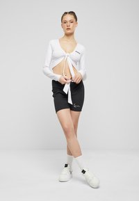 Fehér, hosszú ujjú crop top, elől megkötő részlettel és mély V-nyakkivágással, fekete logós rövidnadrággal és fehér, magas szárú tornacipővel párosítva.