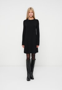 Robe noire à manches longues avec une coupe droite, texture douce, longueur genoux, associée à des bottes noires jusqu'aux genoux avec une finition lisse.