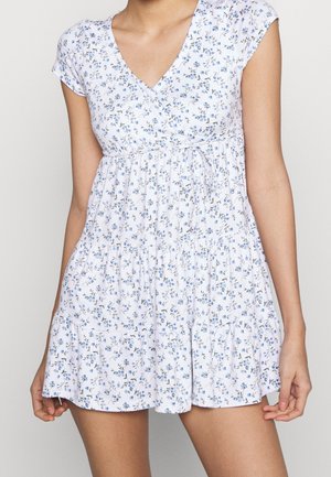 Frau trägt ein kurzes, weißes Blumenkleid mit blauen Blumen, V-Ausschnitt, kurzen Ärmeln und einem Stufenrock mit Kordelzug in der Taille.