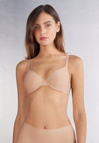 Beige naadloze beha met gewatteerde cups, verstelbare bandjes en een gladde band, met een effen textuur en minimale details.