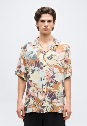 Noor mees seisab, kandes lühikeste varrukatega nööbitavat särki, millel on suur õieprint tuhmides pastelltoonides.