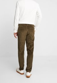 Pantalons en velours côtelé vert olive avec une coupe droite, dotés de deux poches arrière, portés avec une chemise blanche à manches longues et des baskets blanches.