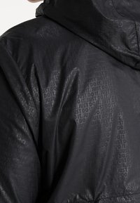 Tissu de veste noire avec un motif répétitif en relief du texte « JUST DO IT » et capuche attachée visible de dos et de côté.