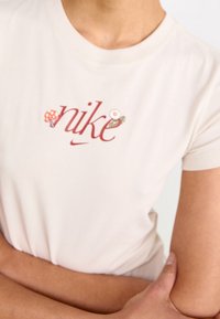 Crème-kleurig Nike T-shirt met rode geborduurde tekst en ontwerpen van paddenstoelen en bloemen. Korte mouwen, zachte stof en een klassieke pasvorm.