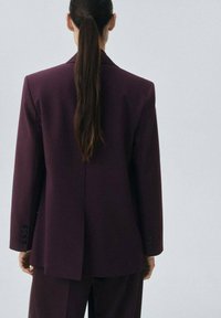 Blazer oversized violet, tissu texturé, fente unique à l'arrière, poignet à quatre boutons, associé à un pantalon assorti, design minimaliste.