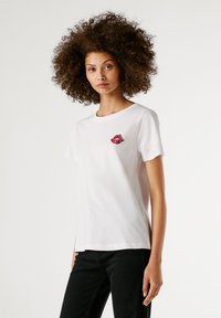 Γυναίκα με σγουρά μαλλιά, ντυμένη με λευκή μπλούζα T-shirt με ένα μικρό λογότυπο σε σχήμα κόκκινου χείλους και μαύρα παντελόνια, που στέκεται μπροστά σε ένα απλό φωτεινό φόντο.