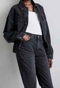 Chaqueta de denim negra con cierre de botones, grandes bolsillos y un corte relajado, combinada con pantalones vaqueros negros de cintura alta. Tela texturizada.