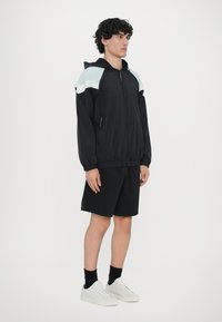 Lahki črni jopič s kapuco, s svetlozelenimi ramenskimi poudarki, z zadrgo žepi, v kombinaciji s črnimi kratkimi hlačami in belimi športnimi copati.