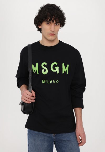 Svart langermet T-skjorte med fet grønn "MSGM" og "MILANO" tekst; løs passform; kombinert med blå jeans og en svart crossover-veske.