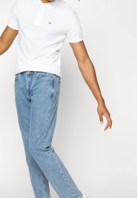 Vit pikétröja med krage, med en liten logotyp. Bärs med ljusblå denimjeans som har en avsmalnande passform och en avslappnad stil.