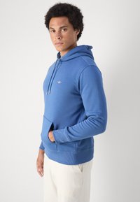 Jonge man met krullend haar die een blauwe hoodie met trekkoorden en witte broek draagt, staat zijwaarts met één hand in de zak.