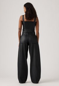 Femme aux cheveux bouclés portant un haut sans manches noir et un pantalon large noir, se tenant de dos devant un fond neutre.