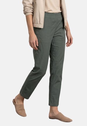 Person, die dunkelgrüne gerade geschnittene Hose, beige Jacke und beige Slipper trägt, steht vor einem schlichten hellen Hintergrund.