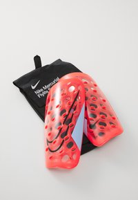 Une paire de protège-tibias Nike Mercurial Flylite rouges avec des logos noirs reposant sur un sac à cordon noir de la marque Nike.
