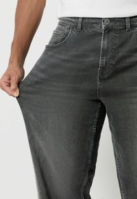 Ciemnoszare dżinsy wykonane z denimu, o prostym kroju, z przednią kieszenią, szlufkami na pasek oraz widocznym przeszyciem wzdłuż szwów. Ręka chwytająca tkaninę.