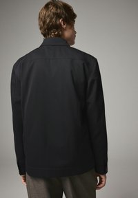 Sort jakke med lige snit, lavet af et struktureret stof. Har krave, knaplukning ved ærmerne og diskrete stikninger på ryggen.