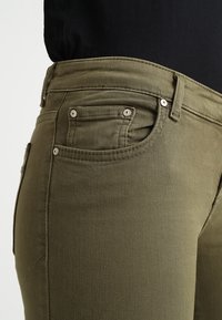 Primer plano de un bolsillo y la cintura de unos pantalones verde oliva con remaches metálicos, llevados con una camiseta negra.