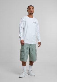 Chemise blanche à manches longues avec un logo imprimé, assortie à un short cargo vert clair avec des poches latérales, et des baskets blanches avec des chaussettes jusqu'à la cheville.