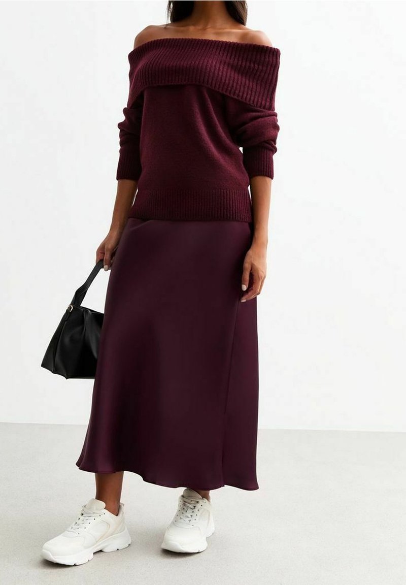 Femme portant un pull bordeaux à épaules dénudées et une jupe midi assortie, tenant un sac à main noir et portant des baskets blanches, debout sur un sol gris.