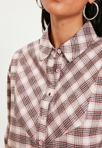 Camicia a scacchi rosa e nera con colletto classico, chiusura con bottoni e una texture di tessuto morbido. Caratterizzata da bottoni rosa e un dettaglio della tasca.
