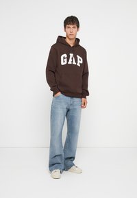 Brown hoodie with large white "GAP" logo, front pocket, paired with light blue wide-leg jeans and white sneakers on a plain background.