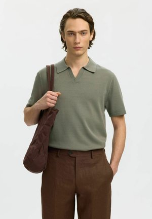 Jeune homme aux cheveux bruns portant un polo vert olive à manches courtes et un pantalon marron, avec un sac à bandoulière marron assorti sur fond blanc.
