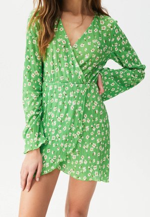 Femme portant une robe portefeuille verte à manches longues avec un imprimé floral blanc et un ourlet à volants, debout avec une main sur la hanche.