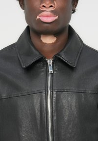 Black leather jacket with a spread collar, front zipper, textured finish, and seamless shoulder design. Features a minimalistic style.