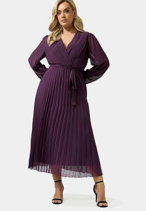 Femme portant une robe portefeuille plissée à manches longues, violet foncé, avec une ceinture à la taille et des sandales noires à talons hauts, debout, les mains sur les hanches.