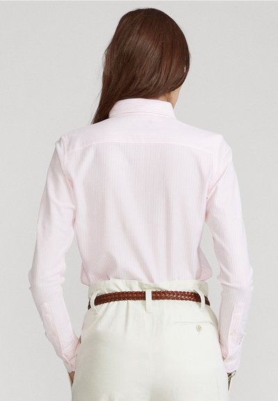 Femme avec de longs cheveux bruns portant une chemise rayée rose pâle rentrée dans un pantalon beige taille haute, avec une ceinture tressée marron.