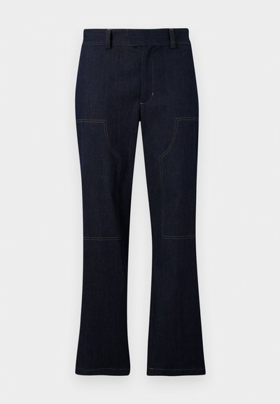 Dunkelblaue, schmal geschnittene Jeans mit hohem Bund, sichtbaren Nähten und zwei großen Front-Patch-Taschen. Weiche Textur mit leichtem Stretch.