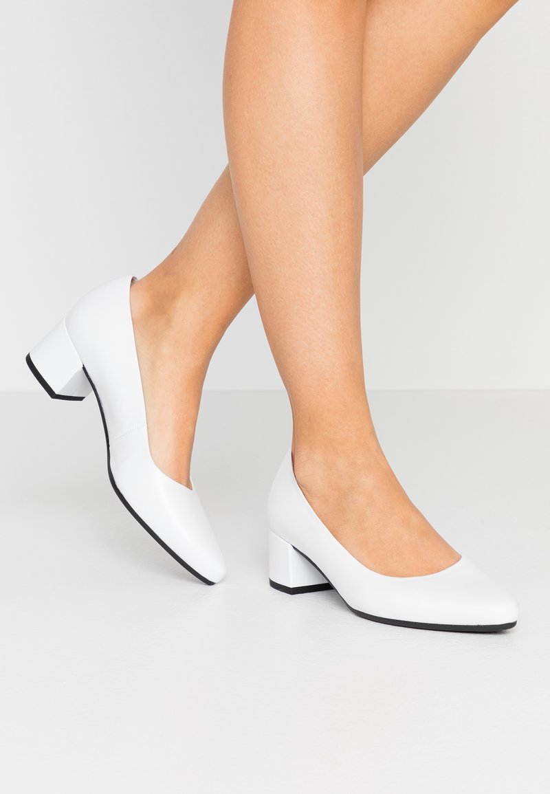 Jambes nues portant des escarpins à talons carrés en cuir blanc avec bouts arrondis sur un sol et un arrière-plan gris clair.