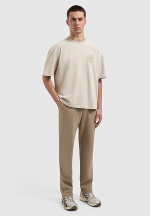 Young man standing with hands in pockets wearing beige oversized T-shirt, taupe pants, and silver sneakers on plain light background.