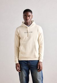 Felpa beige con cappuccio e tasca frontale, con la scritta "Marc O'Polo DENIM" in un leggero lettering scuro. Tessuto morbido e polsini a costine.