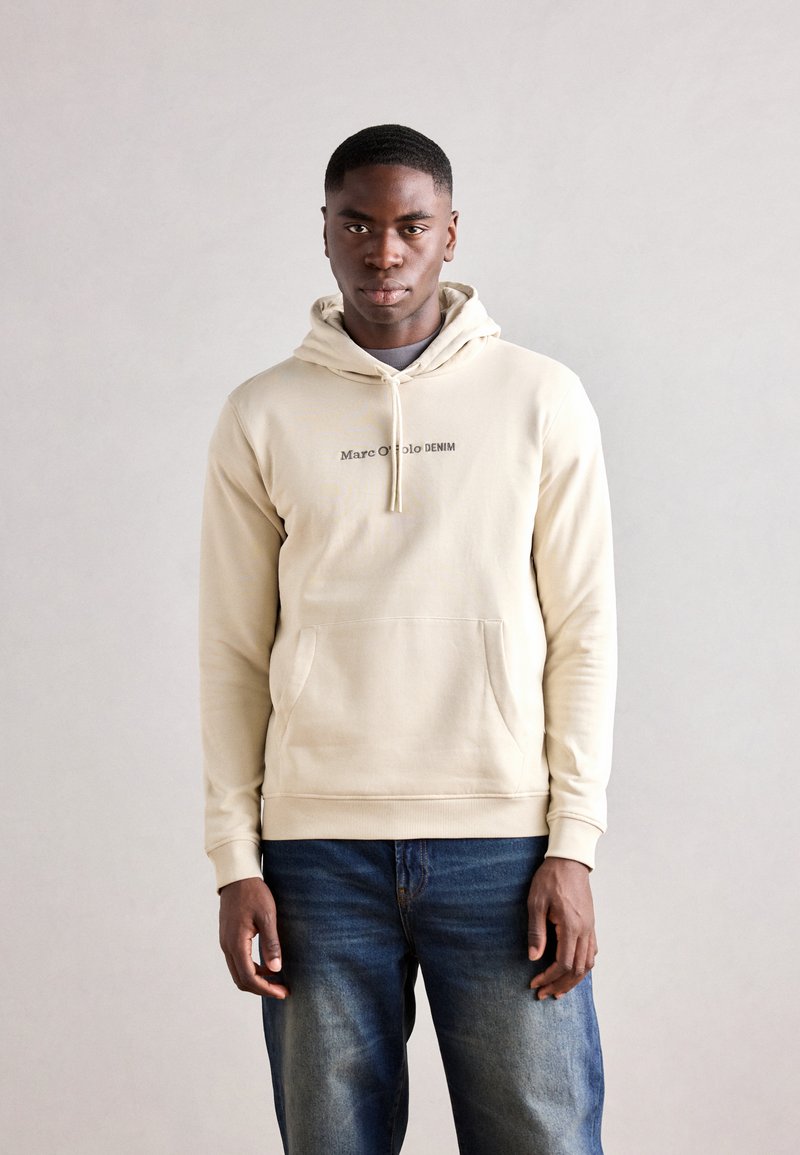Felpa beige con cappuccio e tasca frontale, con la scritta "Marc O'Polo DENIM" in un leggero lettering scuro. Tessuto morbido e polsini a costine.