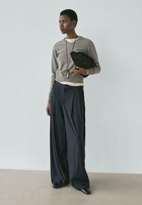 Grauer Strickpullover, marineblaue Hosen mit weitem Bein, schwarze Clutch, lange Anhänger-Halskette. Glatte Texturen, neutrale Farben, entspanntes Design.